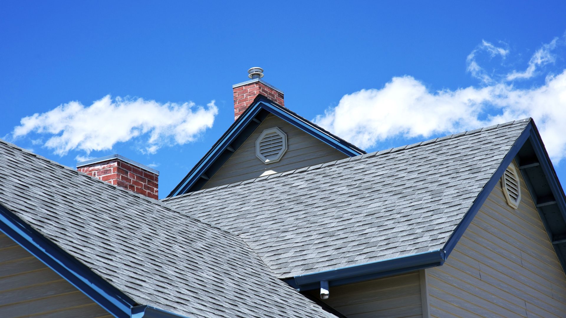 Gray shingled roof with brick chimneys against bright blue sky with clouds